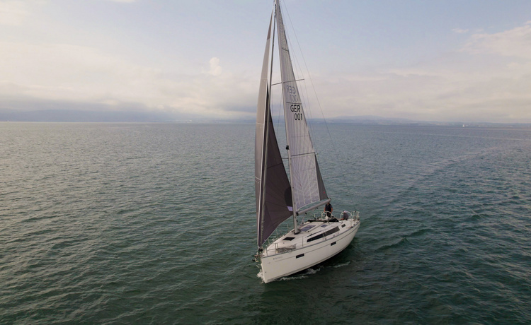 Zwei Personen steuern ein modernes Segelboot über ruhiges Wasser, die Kamera zeigt eine leicht schräge Frontansicht mit sichtbarer Bugwelle.