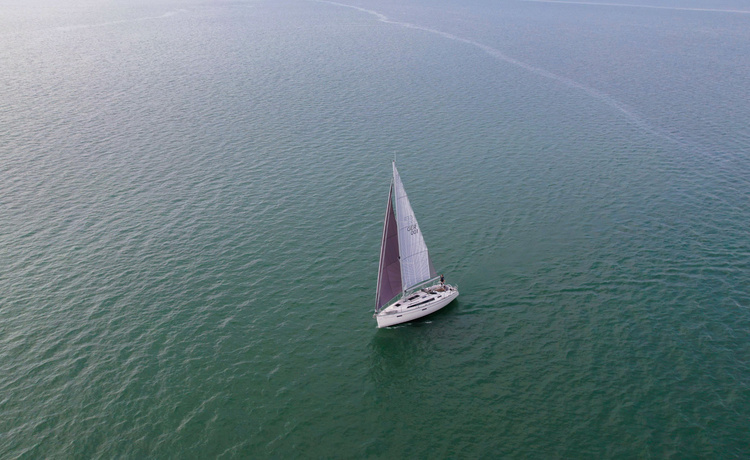 Luftaufnahme einer weißen Segelyacht mit dunklem Großsegel auf ruhiger, grüner Wasserfläche, keine Küste in Sicht.