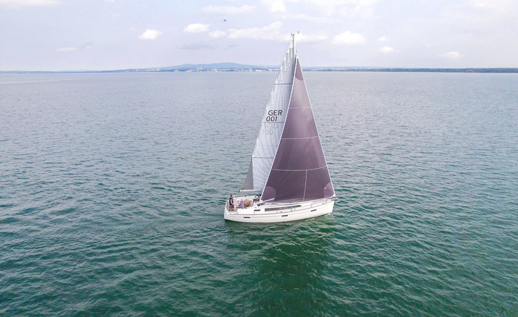 Segelboot mit dunklem Großsegel und der Kennung „GER 001“ fährt auf ruhigem Wasser, im Hintergrund ist die entfernte Küste sichtbar.