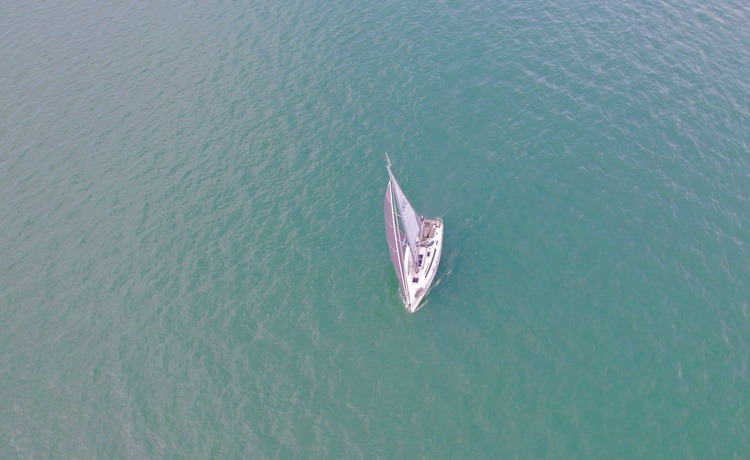 Von oben aufgenommenes Segelboot mit auffälligem Segel in hellem Wasser, das eine leichte Kursänderung vollzieht.