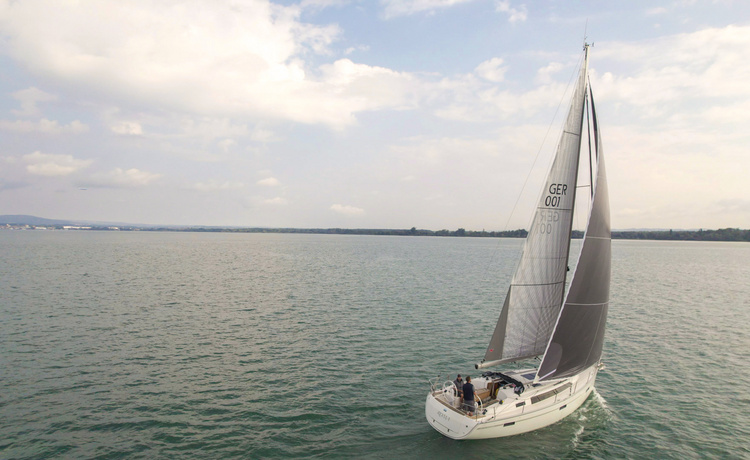 Modernes Segelboot mit zwei Personen an Bord fährt nahe der Uferlinie, im Hintergrund flache Hügellandschaft und bewölkter Himmel.